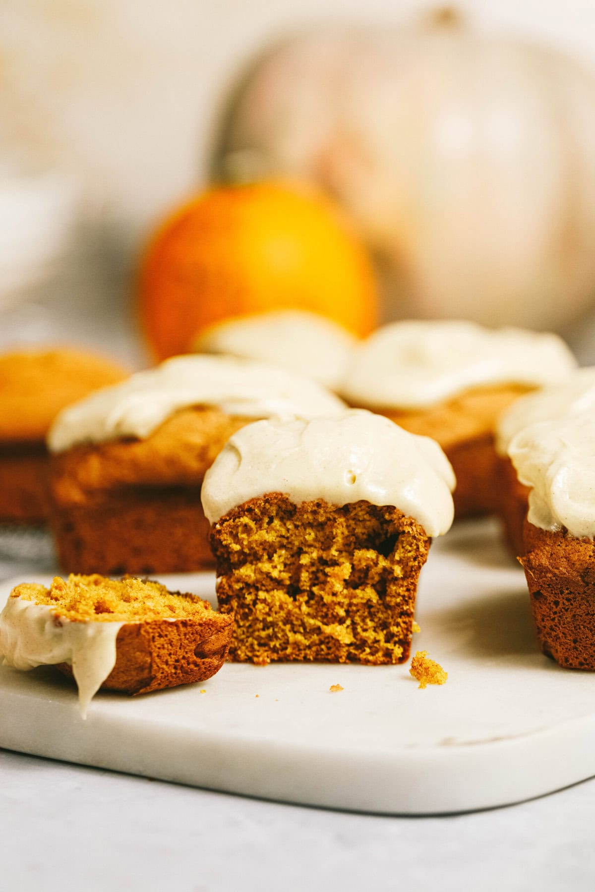 Close-up of several pumpkin muffins topped with white frosting, one partially eaten, on a white surface with a pumpkin and an orange fruit in the background.