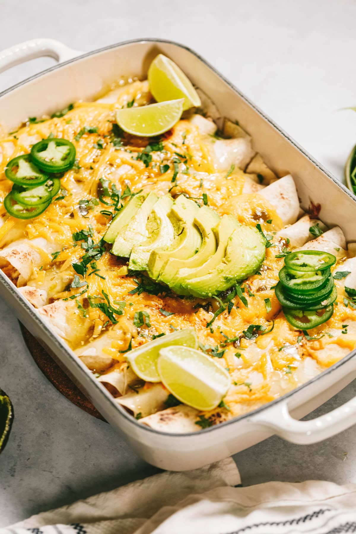 A baking dish filled with enchiladas topped with melted cheese, sliced avocado, lime wedges, and jalapeño slices on a light-colored surface.