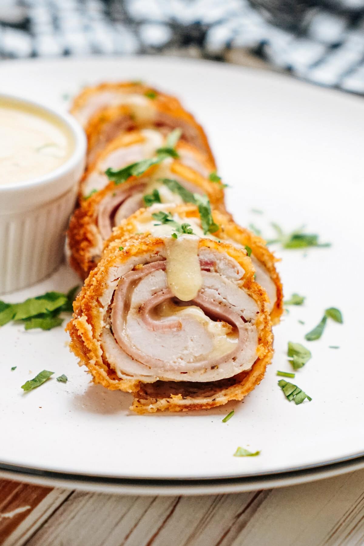 Sliced chicken cordon bleu rolled with ham and cheese, garnished with chopped herbs, served with a side of creamy dipping sauce on a white plate.
